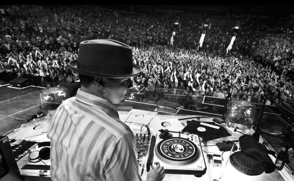 A black and white photo of a DJ wearing a fedora in front of turntables at a concert.