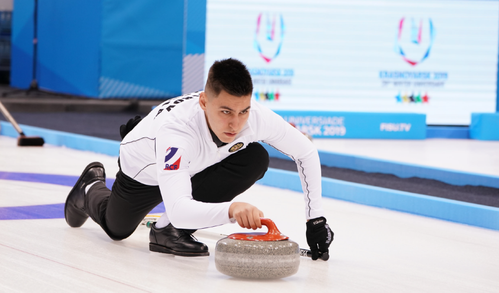 A male curler slides on ice, pushing a curling stone.