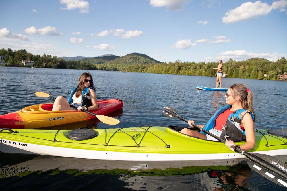 Two women kayak and another paddles a SUP on Mirror Lake, all of which can be found at Lake Placid boat rentals.