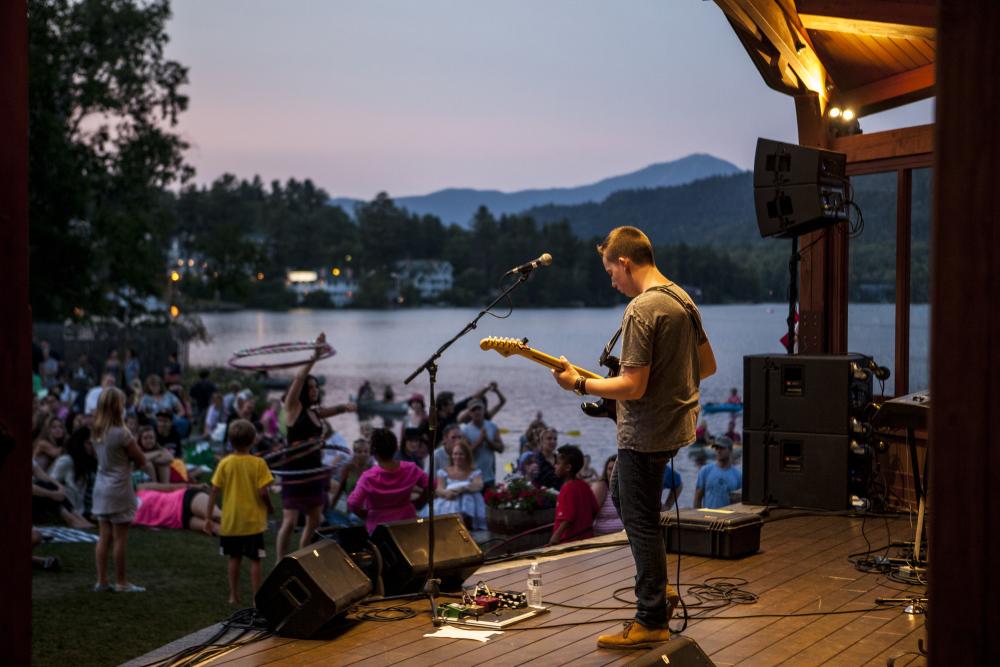 A man playing guitar on a stage next to a lake with people dancing in the background.