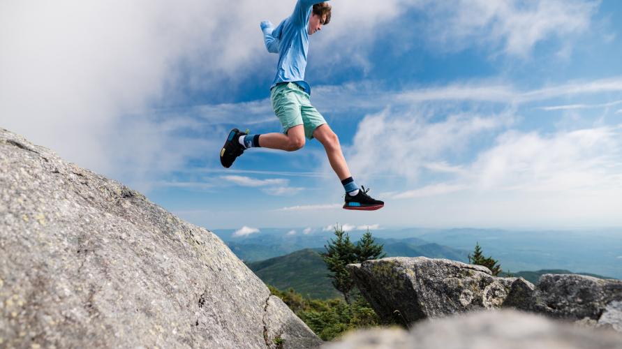 A young boy jumps from a rock to another rock on top of a mountain. 