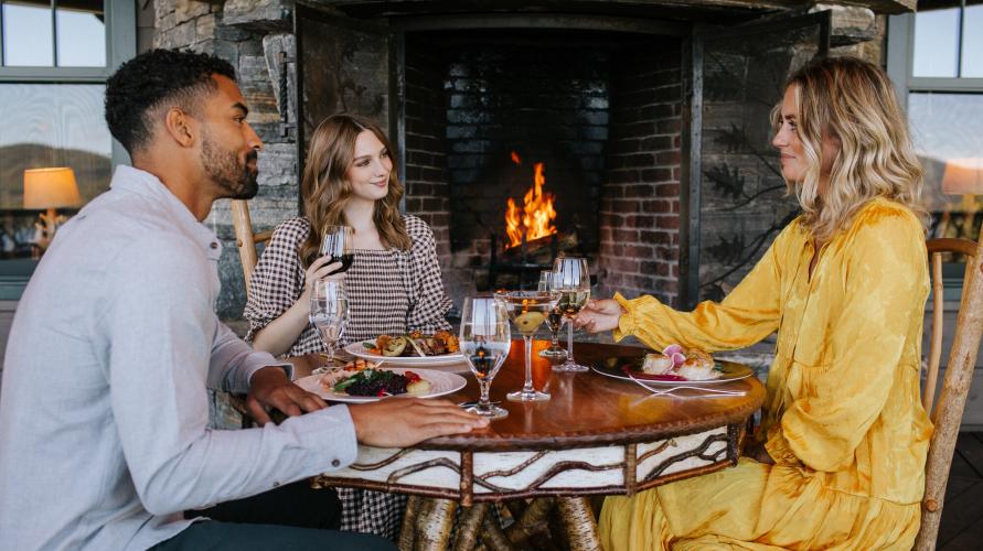 Two women in dresses and a man in a collared shirt and sweater eat breakfast by a stone fireplace