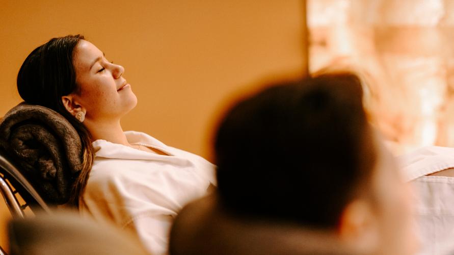 Woman relaxes reclined in robe in warmly lit salt room next to a blurred subject.