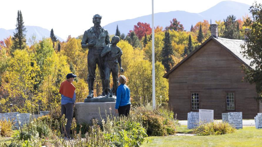 Two people stand at the base of John Brown's statue in Lake Placid with his farm in the background