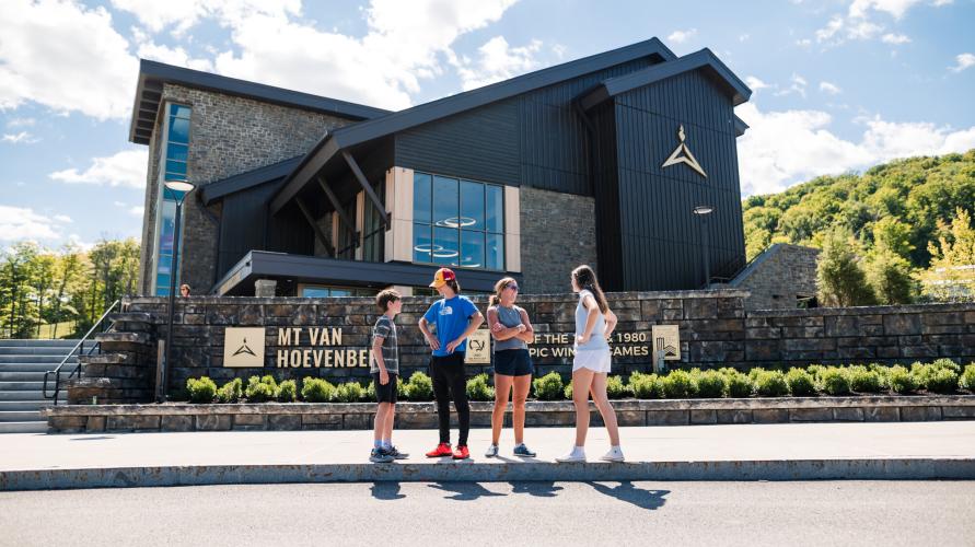 Three teens and a mother stand in front of the Mt Van Hoevenberg Lodge.