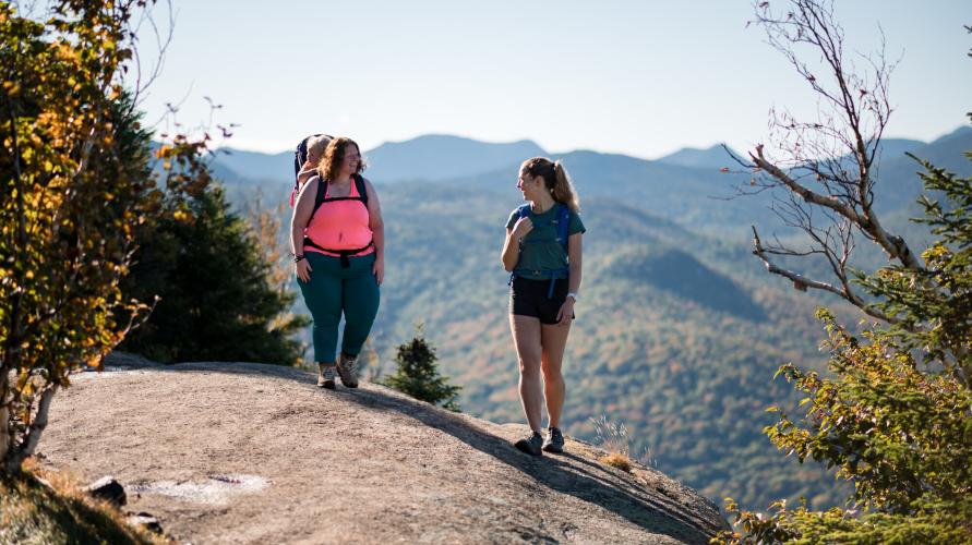 Two women, one with a baby carrier, stand atop a mountain summit