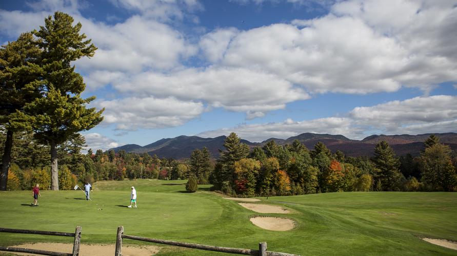 A bright green golf course is framed by fall foliage and a blue, cloud-specked sky on a sunny day.