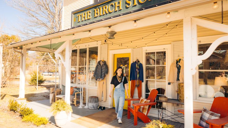 Young woman in overalls walks out of boutique on porch filled with merchandise