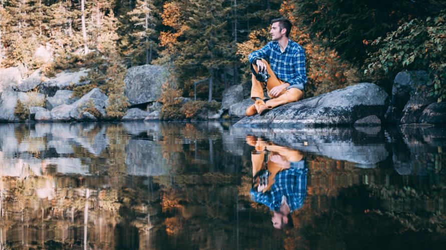 Man sits on rock with camera next to still pond where he is perfectly reflected