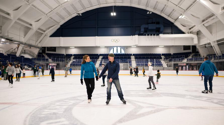 Couple ice skates on indoor rink with other people skating around them