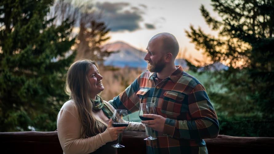 A man and women looking at each other and drinking wine with a picturesque background behind them.