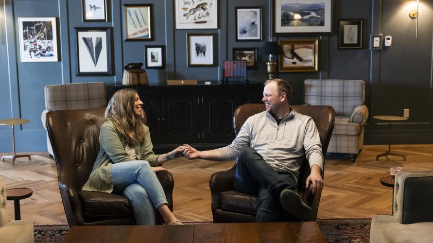 couple sit in two leather chairs in dark blue lounge with lots of photos holding hands