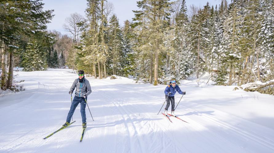 A man and woman ski on a wide snowy path on a sunny day. 