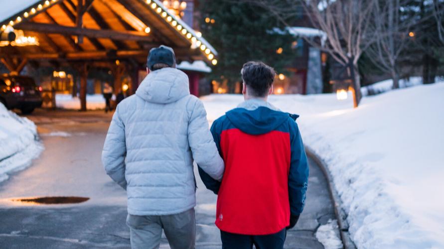 Two men in winter jackets walk to a warmly lit lodge with a log car port. 