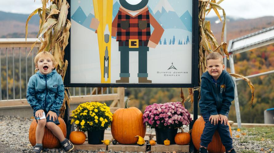 Two kids laugh in front of a sign with pumpkins and mums.