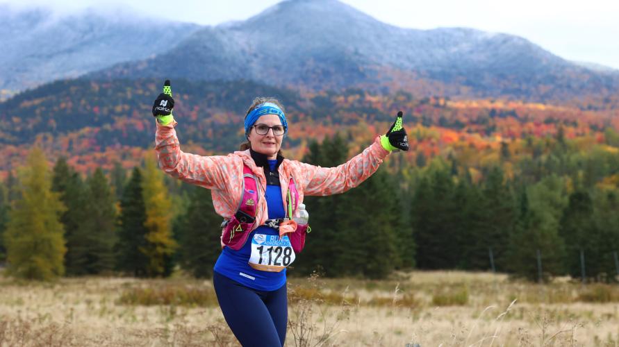 A woman gives two thumbs up while running in front of fall mountains