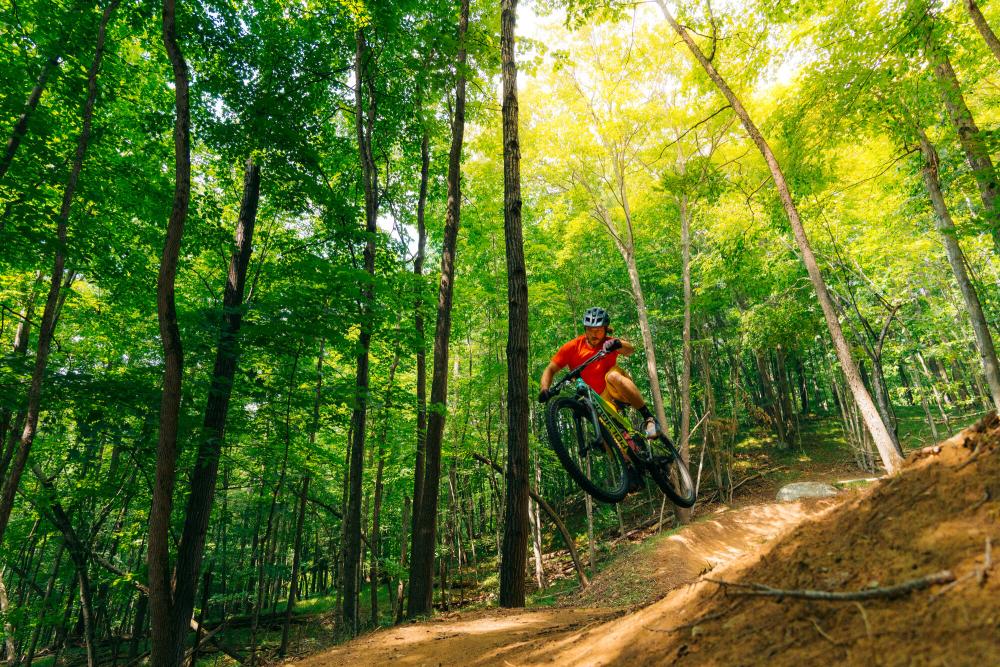 A man in a red t shirt makes a jump on a mountain biking trail.