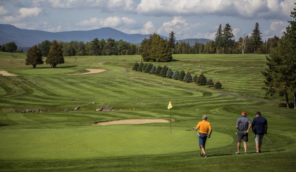 Three ment walk to the flag in a golf course with mountains in the background.