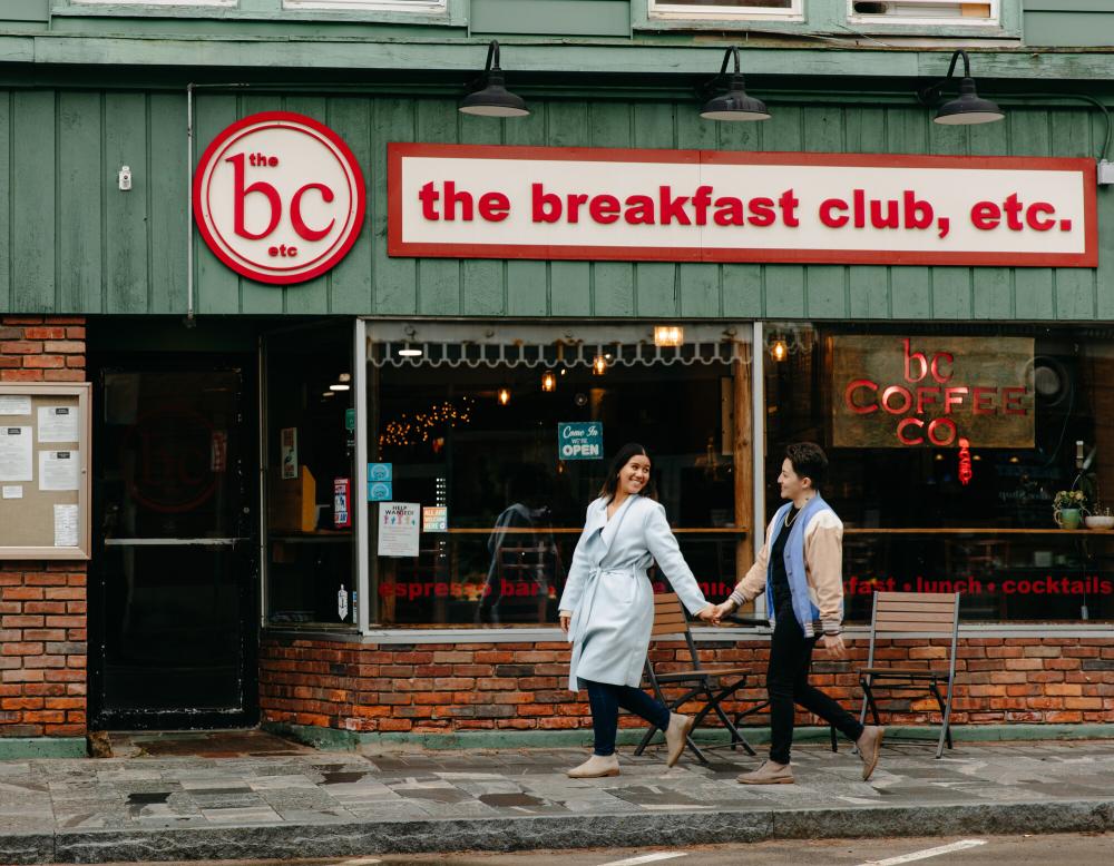 Two woman hold hands, one looking back over her shoulder at the other, walking toward the door of Breakfast Club restaurant