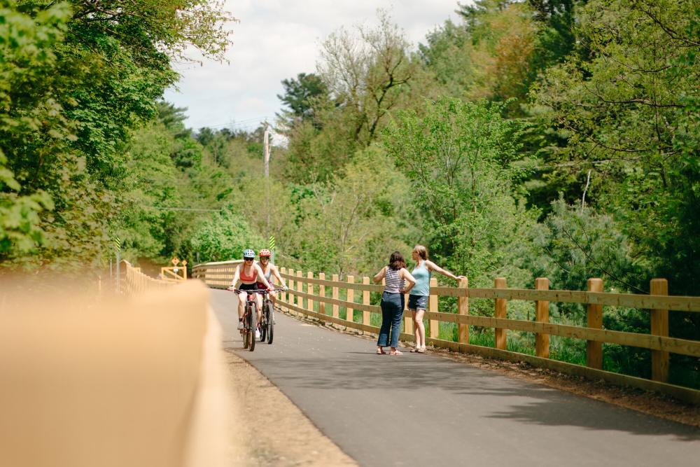 Two women stand on rail trail while bikers pass