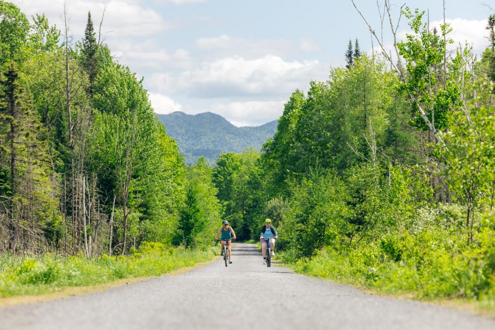 Two bikers ride side by side on rail trail with mountains in background