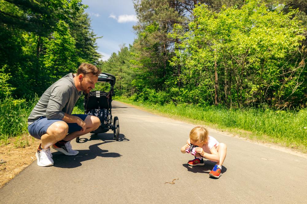 Father with stroller crouches next to small daughter crouched on rail trail looking at snake through binoculars