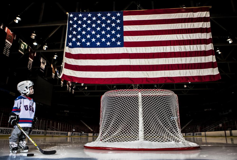 A small child in hockey gear looks upward at an American flag over a rink.