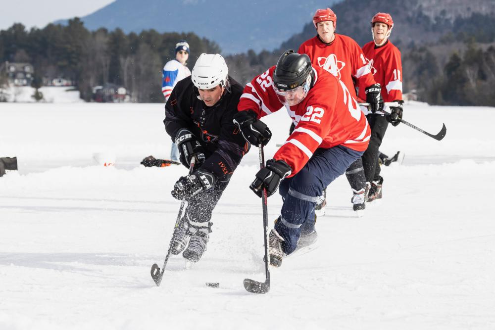 Two hockey players fight for control of the puck on a frozen lake on a sunny day.