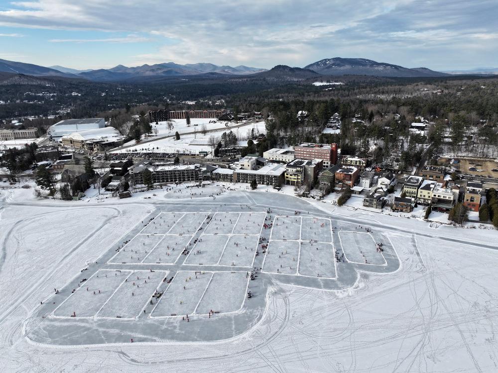 An aerial view of a frozen lake, divided into numerous pond hockey rinks. A small town overlooks the lake.