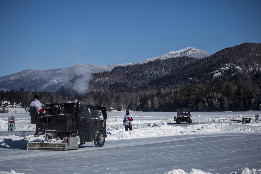 A zamboni driver sweeps across an outdoor rink with snowy mountains in the background.