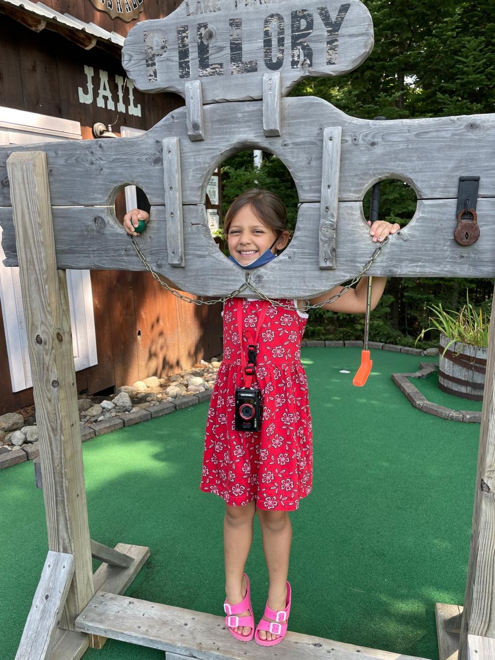 No children were arrested or harmed in this round of golf. A small girl in a red dress poses in a wooden pillory on a mini golf course