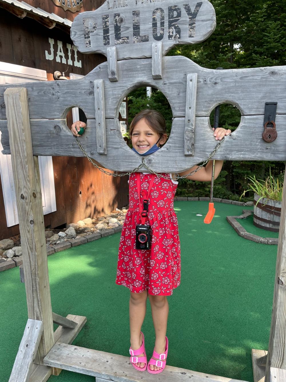 A small girl in a red dress poses in a wooden pillory on a mini golf course