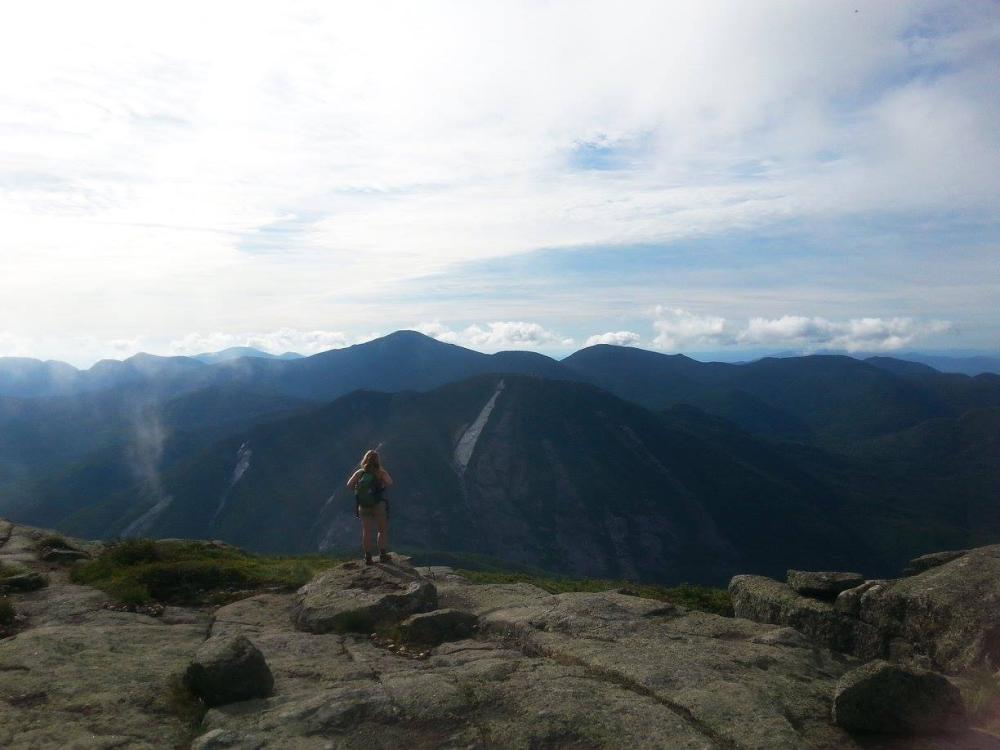 A woman with hiking backpack stands admiring the view in the High Peaks.