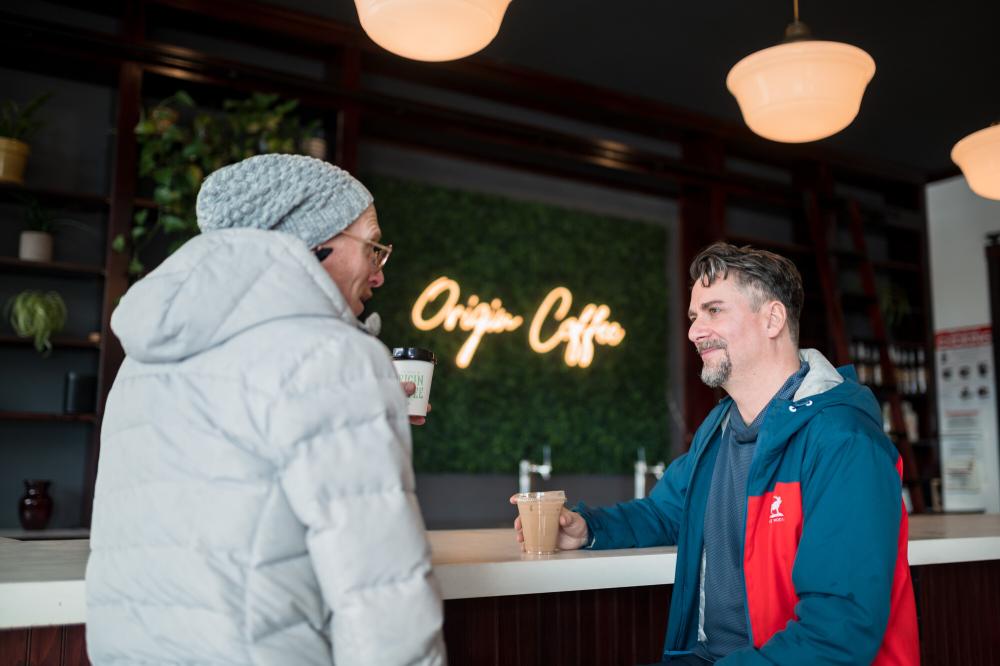 Two men share coffee together at a coffee bar with grass decor wall in the back.