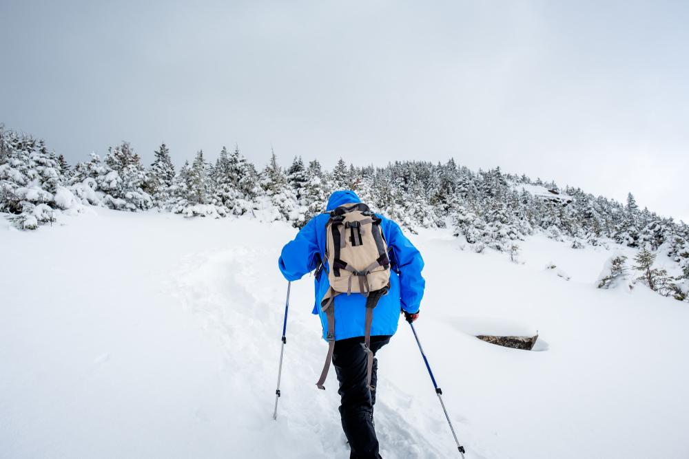 A person climbs a white snowy mountain with outdoor winter gear on and walking sticks.