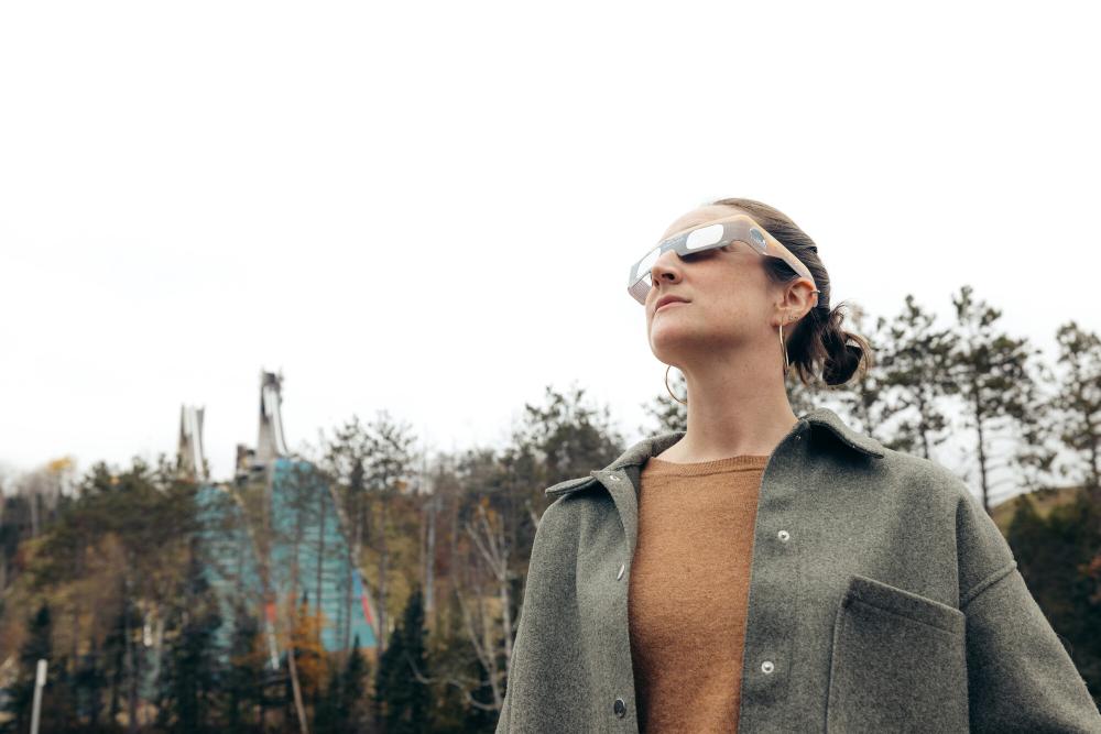 A woman wearing eclipse glasses gazes up at the sky with ski jumps in the background.