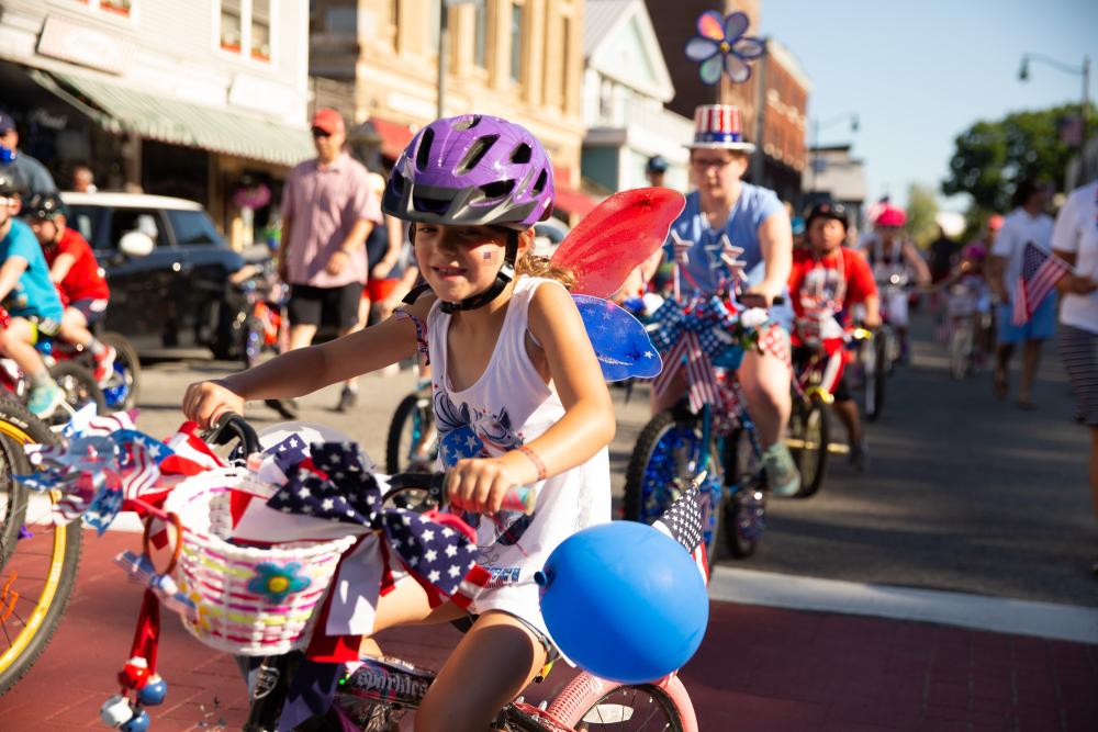A young girl decorated in July 4th colors and ribbons rides a bike in a parade.