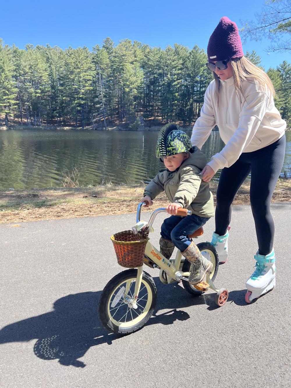 A woman on rollerblades guides a small child on a training bicycle.