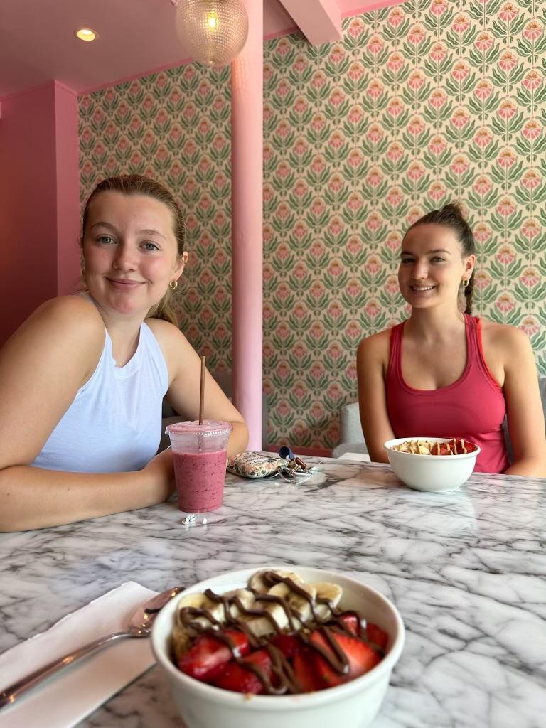 Two young women smile over a light lunch in a small cafe with pink and green decor.