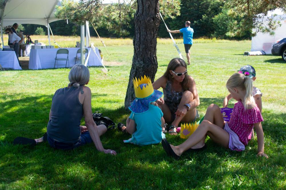 A woman sits under a tree eating snacks with her young kids.