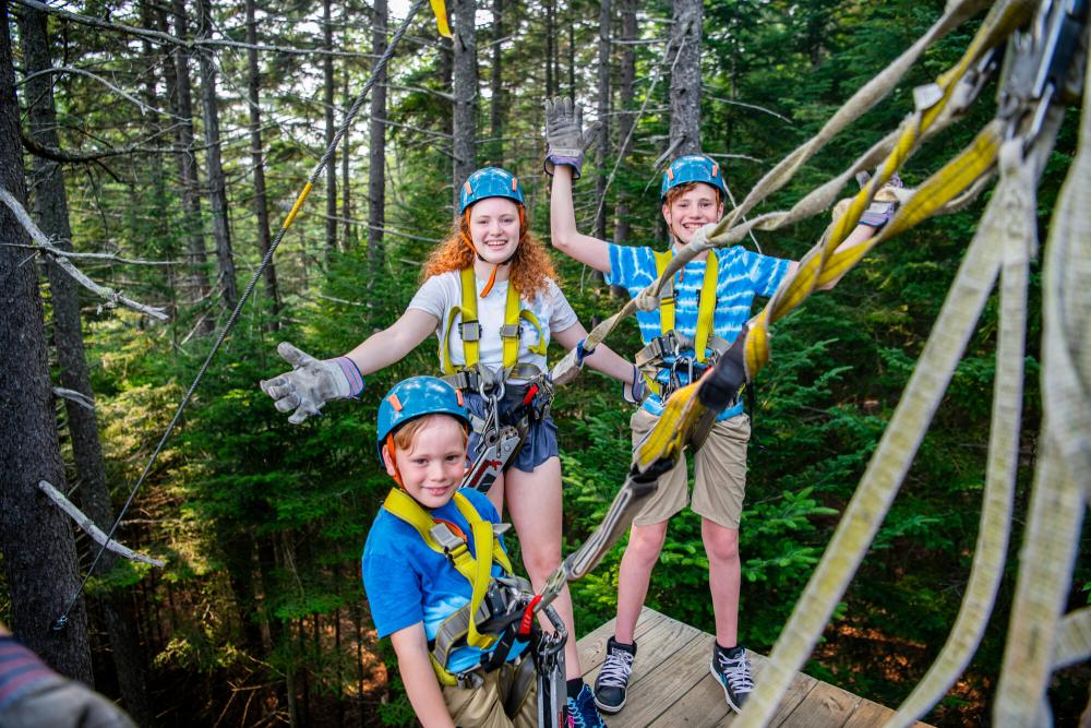 Three children smiling and geared up to go, posing for a picture at the zipline.