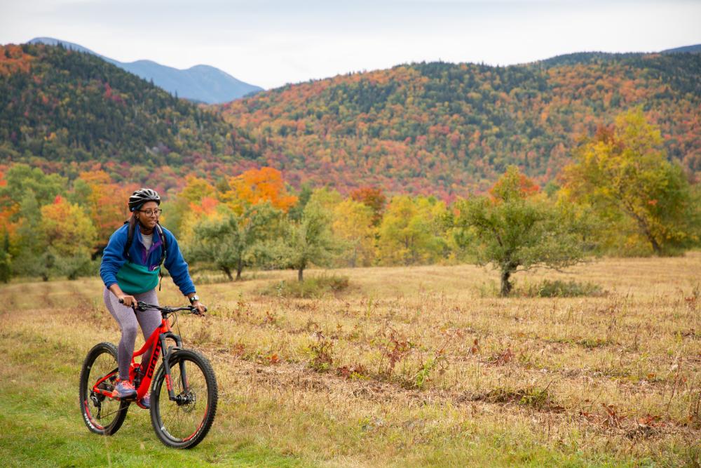 A woman mountain biking on a grassy trail with fall foliage in the background.