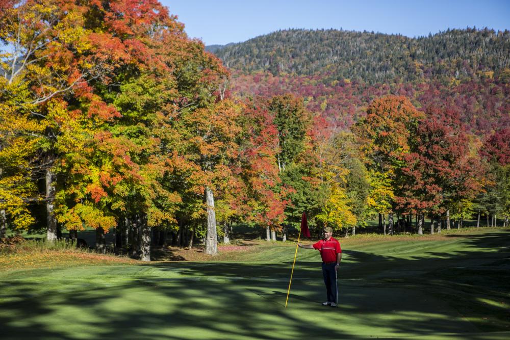 A man standing on the golf course holding a flag and smiling with beautiful fall leaves surrounding him.