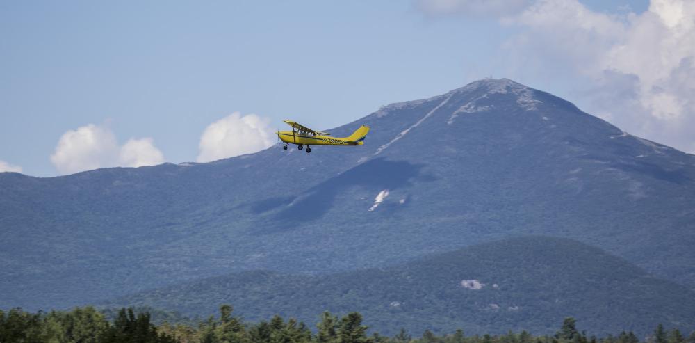 A yellow airplane in flight with mountains in the background.