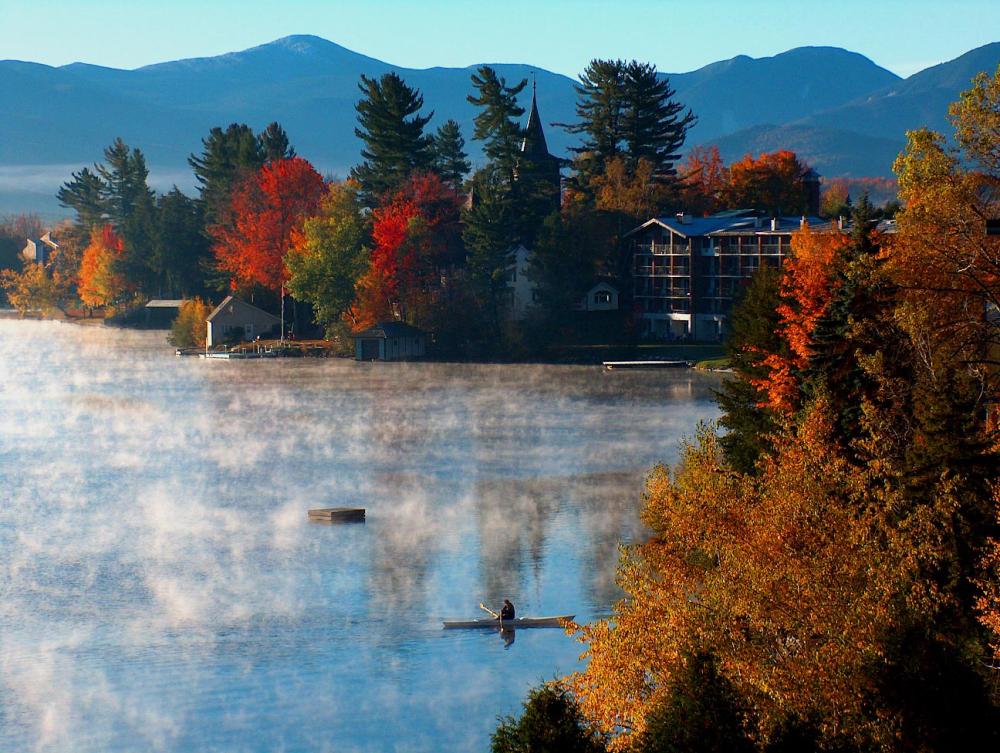 A person is paddling a canoe on Mirror Lake with morning mist coming off the water, fall trees surrounding them, and mountain views in the background.