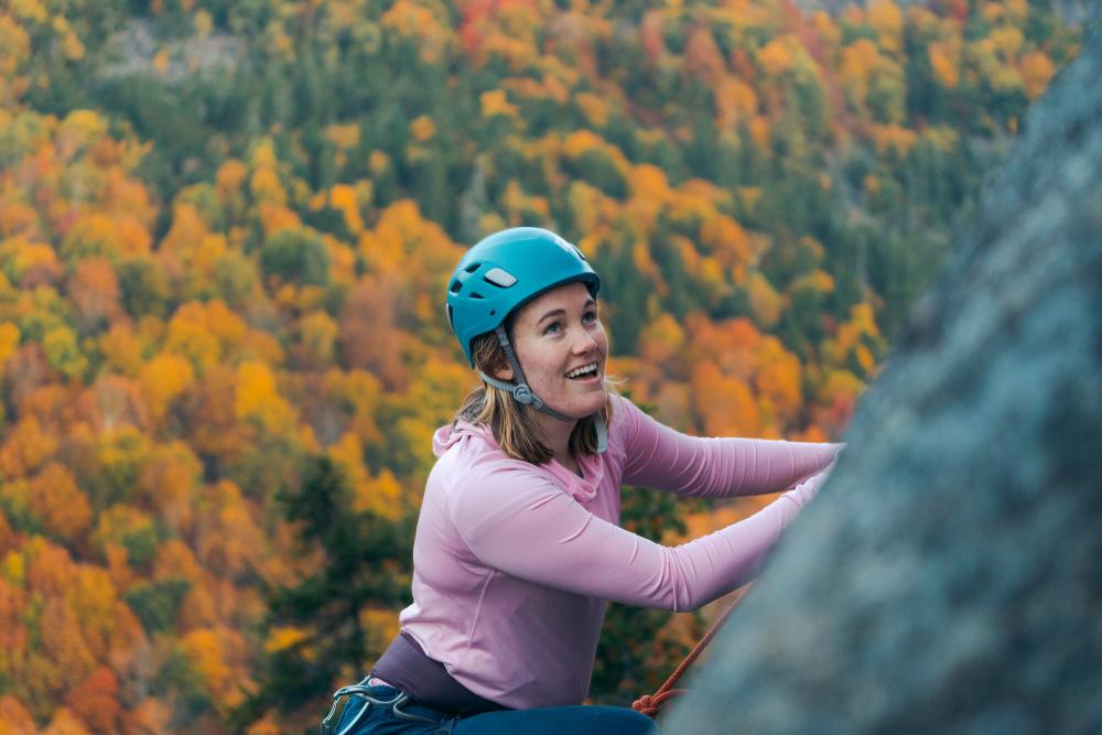 A woman smiling while rock climbing with an aerial view of the fall foliage below her.