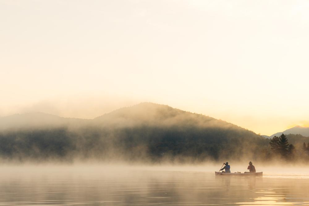 A canoe with two paddlers floats on mist-covered lake with mountain in background