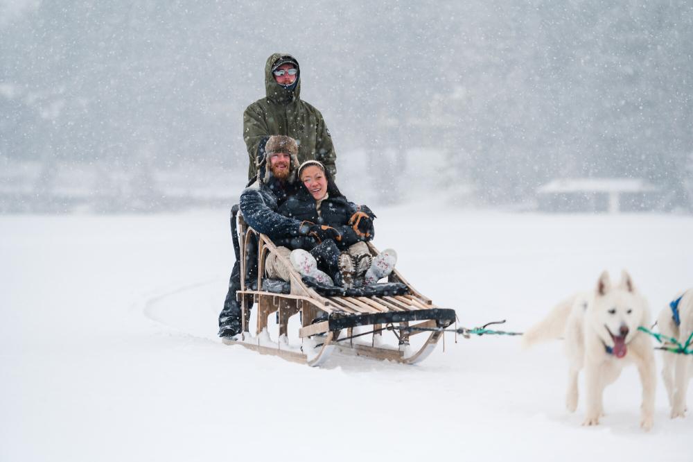 Man and woman sit in sled pulled over frozen lake by dog team