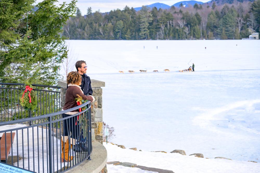 Man and woman stand on patio overlooking frozen lake with dog sleds and distant mountains