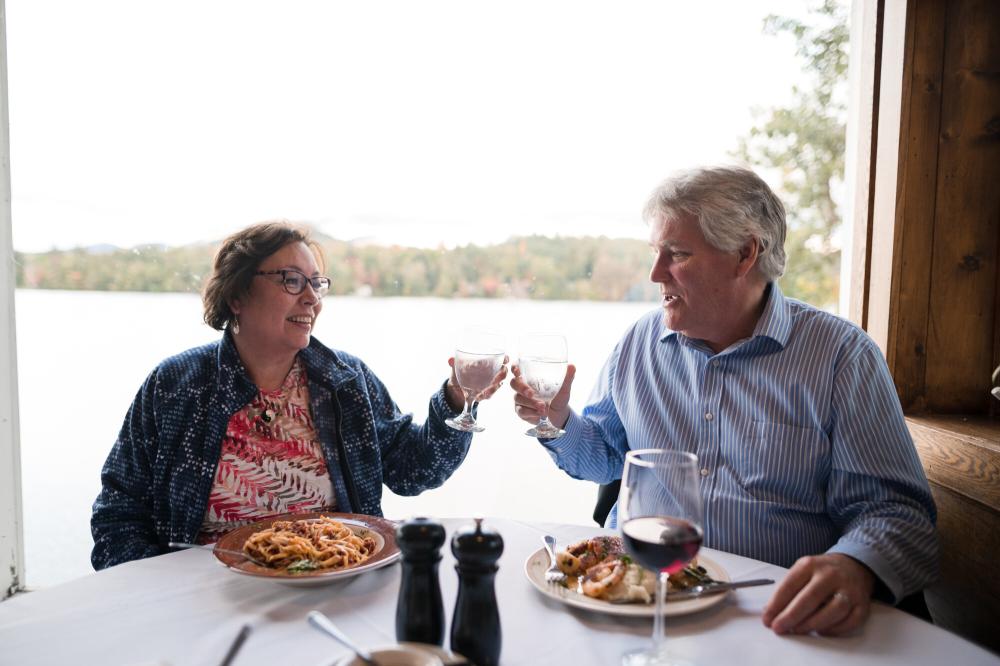 A couple clinking their beverages while dining at Jimmy's 21, seated in front a window with views of Mirror Lake.